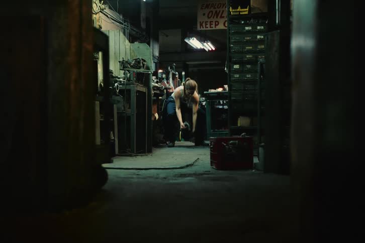 Female car mechanic wearing a white shirt and dark blue pants lifts up car part at the garage