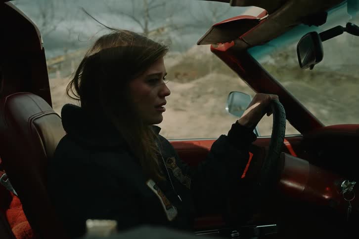A woman with brown hair drives a sports car down a dirt road tears streaming down her face expressing sadness
