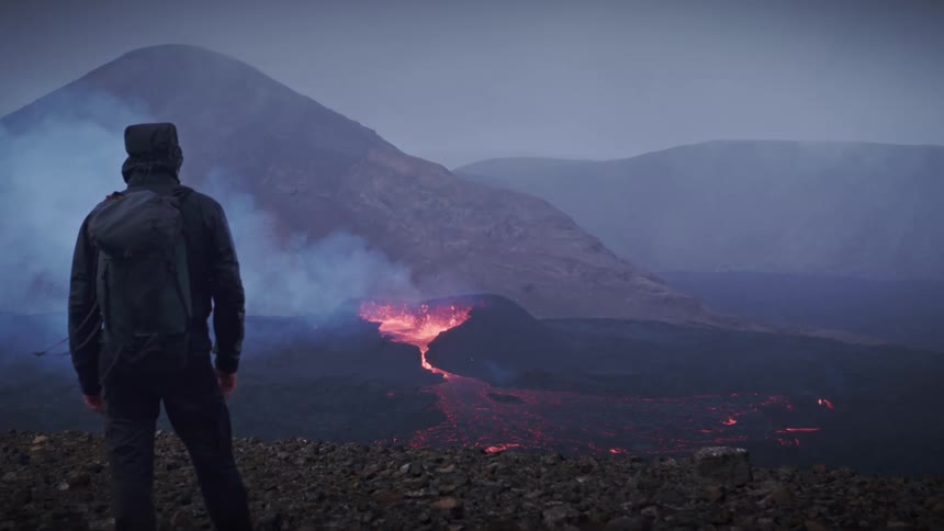 A person standing and watching a volcano erupt with smokes and lava flowing around