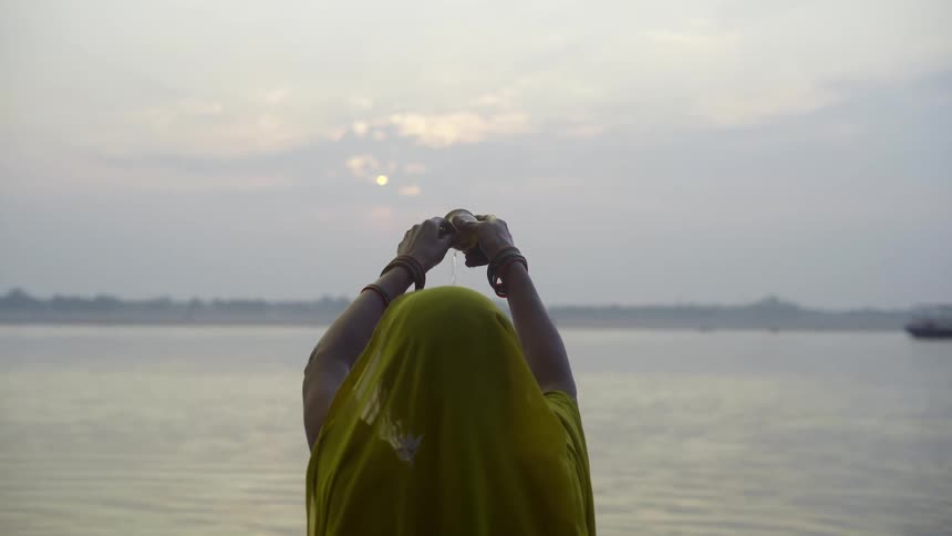 Woman standing on shore raises object toward the sun obscured by clouds