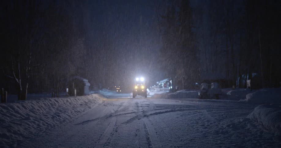 View of a snowcovered road on a night that is snowing and small tractor lighting