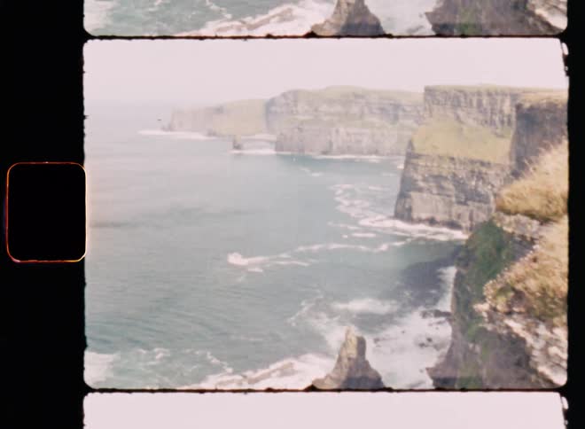 A grassy cliff overlooks rocks and waves crashing at its base