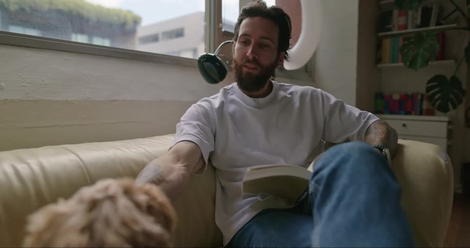 A man sitting on a couch reading a book while petting a dog