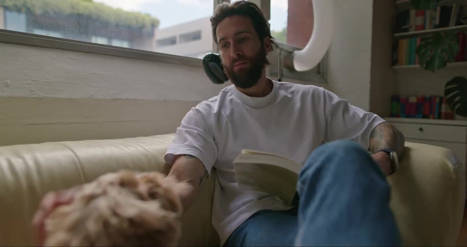Man stops reading his book to pet and talk to his dog while sitting on a leather couch in his living room