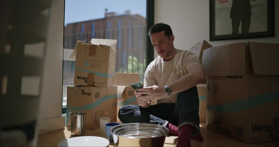 Man sitting on the wooden floor next to a glass window surrounded by boxes scrolling through his phone and drinking from a cup