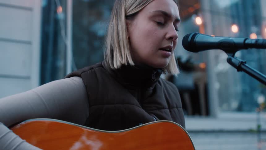 A lady singing and playing guitar outside a building during the day