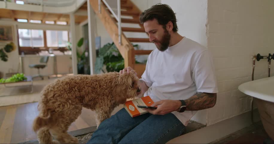 A man with a book in his lap checks his phone while petting his dog