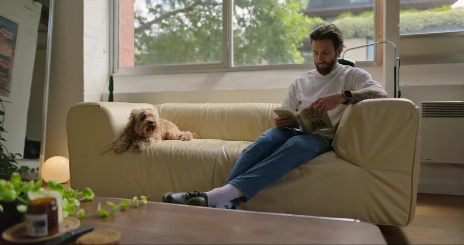 A man is sitting on a sofa indoors while reading a book in the daytime