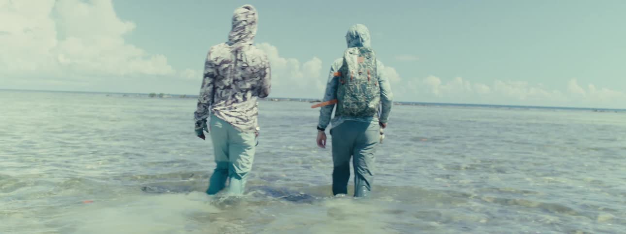 Two men walking inside the sea at midday holding fishing rods