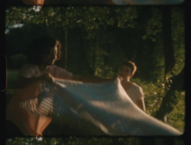 Man laying a towel on the grass by a lake and sitting down with his girlfriend