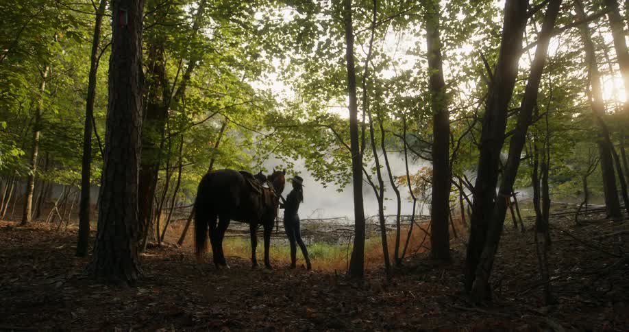 Woman standing beside her horse in the forest caressing it in front of a lake