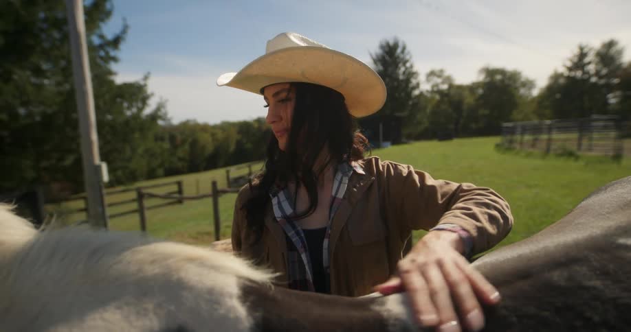 A lady wearing cowboy hat rubs the back of a horse on a farm during the day