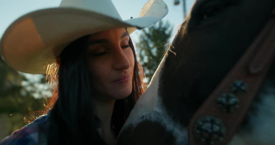 A woman with dark hair and a cowboy hat gazes affectionately at a horse while gently petting it