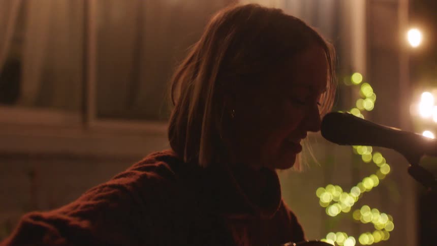 Woman sitting behind an illuminated house playing the guitar and singing into a microphone at night