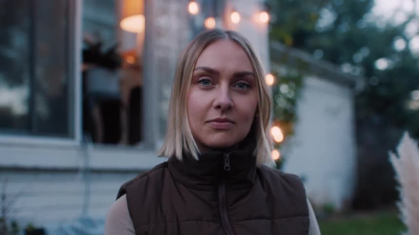 A blonde woman wearing a brown jacket seated in the backyard in front of the window