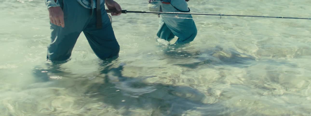 Two men in jackets and caps walking in the sea with fishing rods