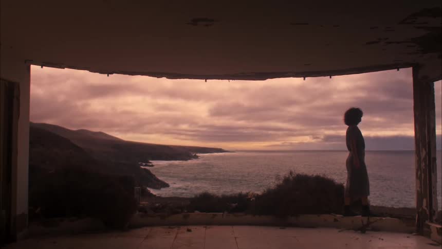 Woman walking on a cliff next to a destroyed house facing the sea in a purple early morning sky