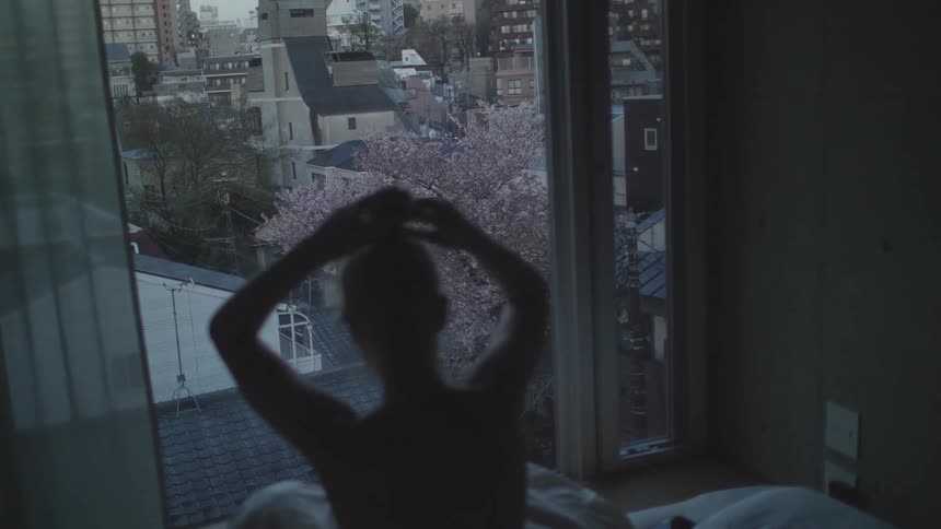 Woman sitting on the bed in front of the open window of her apartment tying her hair and looking at the view of the city buildings ahead