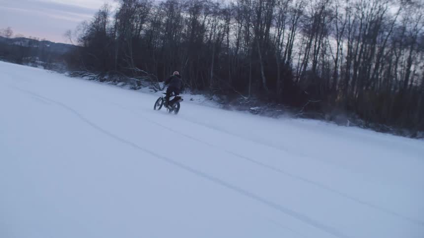 Man on a motorcycle riding through snowcovered terrain outdoors next to an area with numerous leafless trees