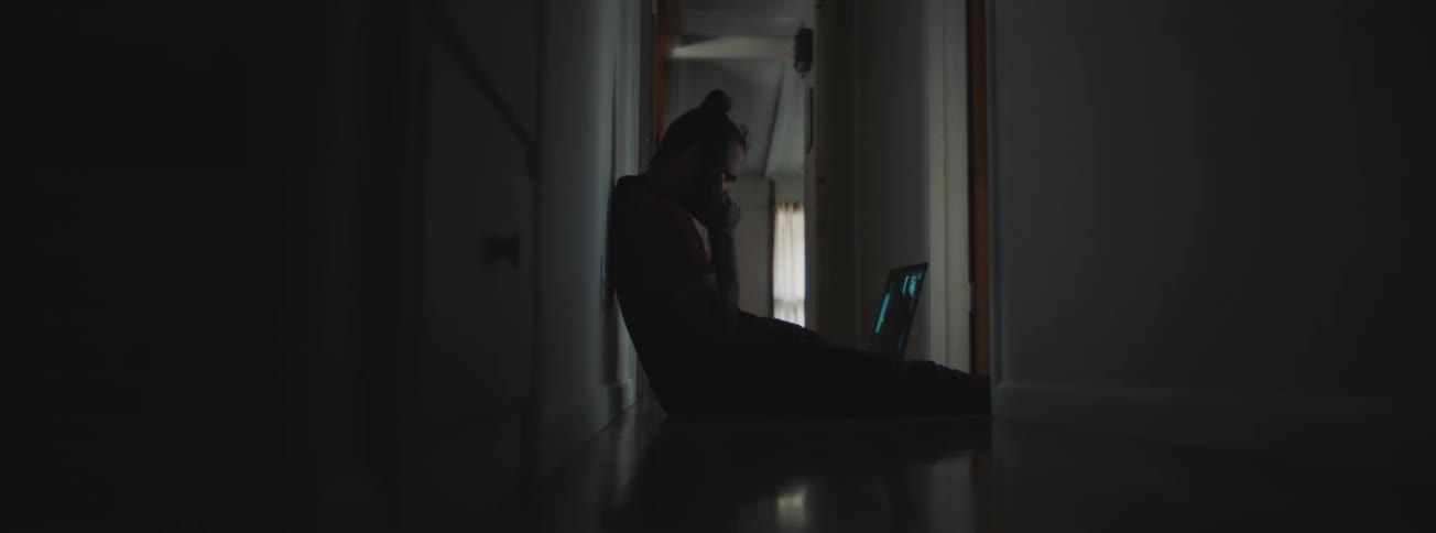 Man sitting on floor in hallway wipes his eyes while viewing video on laptop