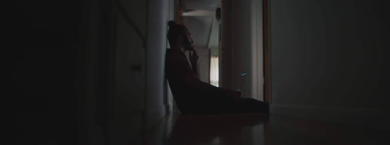 A sad man sitting in the hallway of his house with a laptop on his legs watching family movie