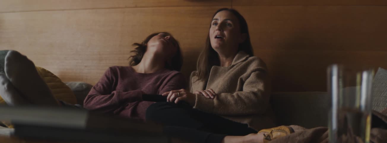A mom sits next to a daughter on the couch and takes the remote as they jokingly argue over the what they are watching