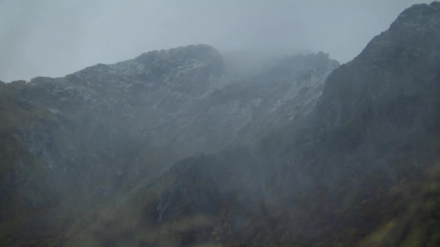 A rocky mountain on a windy day with fog and an overcast sky