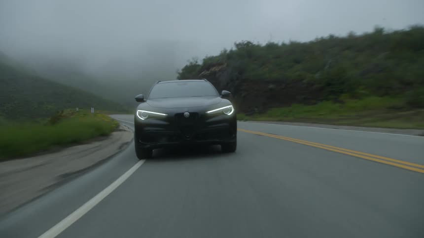 A black car driving on a street with vegetation on the sides and in the sky there is fog