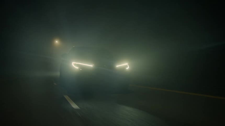 Car with sleek headlights drives down a road through slight fog at night