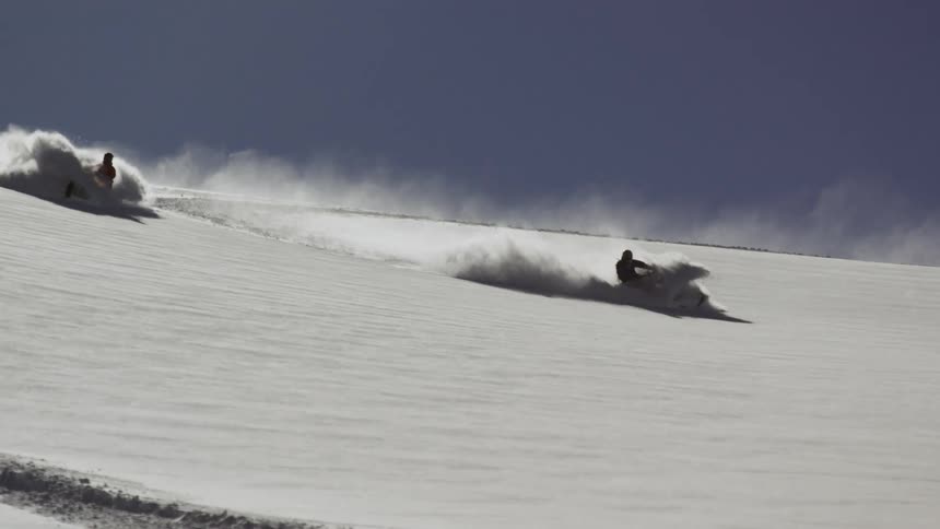 Two snowmobilers ride deep powder downhill on a sunny day