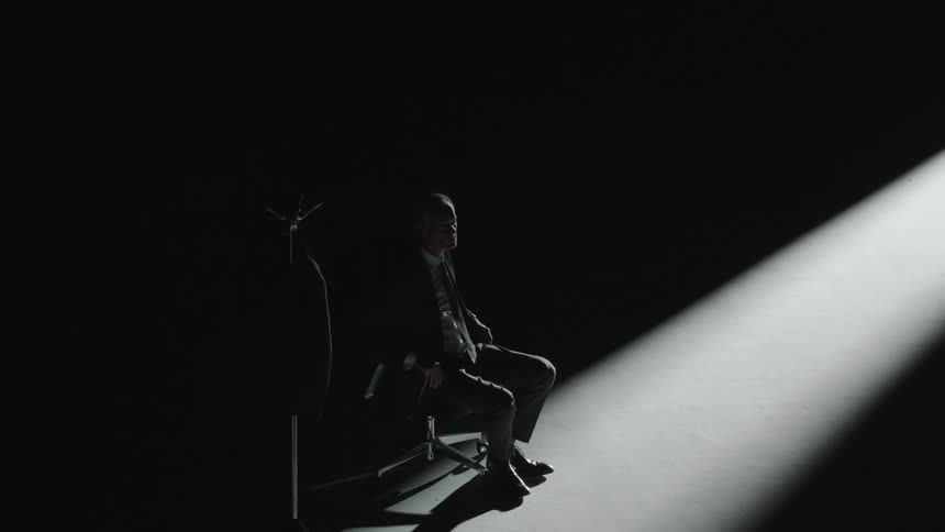 Spotlight partially shining on a man who stands from sitting in a office chair beside a coat rack in an otherwise dark empty room