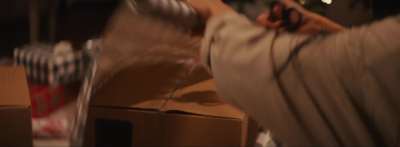 Lady put gift wrapping on a box while seated in a room at night
