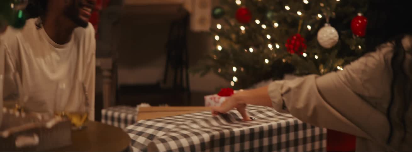 A pair of adults raising a toast with drinks as they wrap christmas gifts seated beside a decorated tree on the living room floor