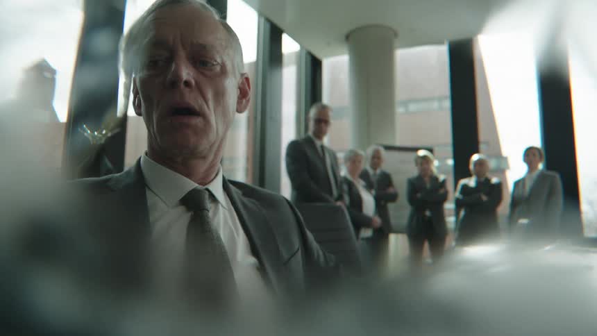Elderly man in a suit sitting and taking a deep breath in desperation with several other people around watching him