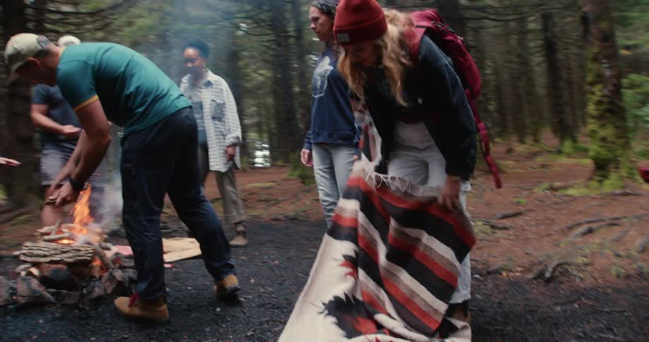 Group of friends around a campfire at the nature adding wood to the fire and laying out a towel on the ground