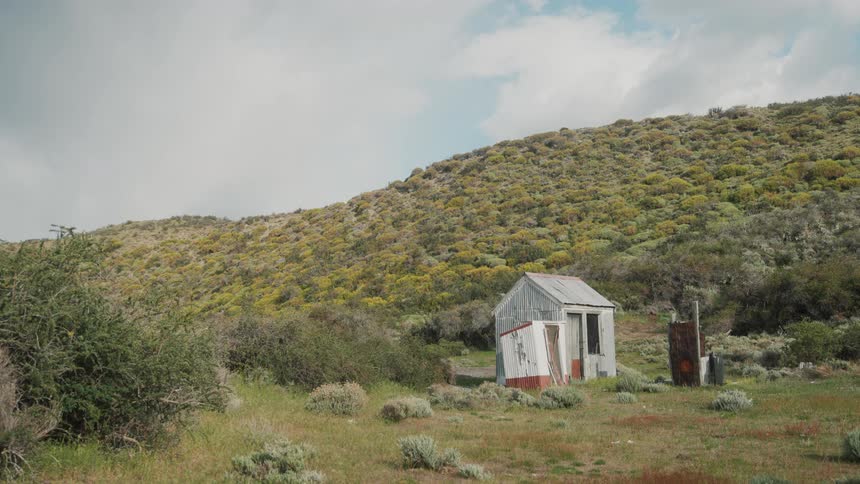 A white shack with connected side building at the foot of some weedfilled hills