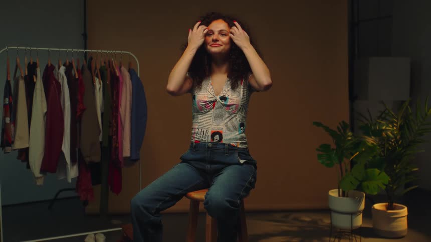 Female teen redress her curly hair while sitting on a stool in a room