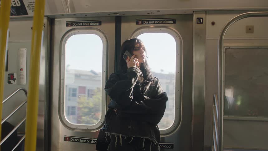 A teenage girl stands in front of the doors of a moving train talking on her phone