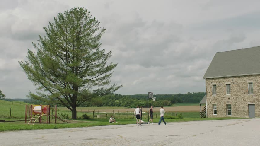 Men on a court next to a field playing basketball on a cloudy day