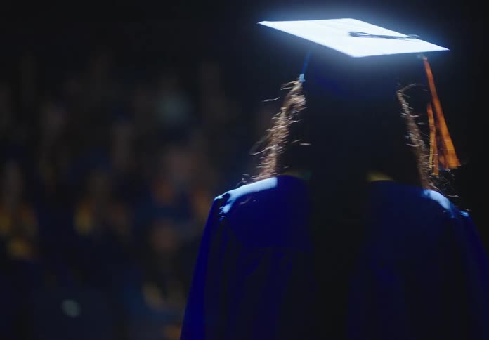 A female wearing a cap and gown shakes a ladys hand and begins to speak to her graduating class