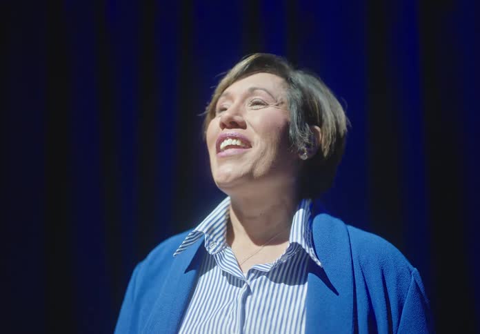 Woman wearing blue striped shirt and sweater stands in spotlight and speaks