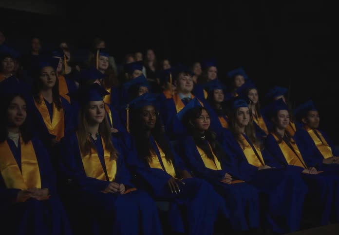 Graduates in blue and gold gowns sitting and two standing up causing applause