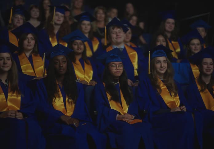 Teenagers in graduation gowns sitting and applauding at a graduation ceremony
