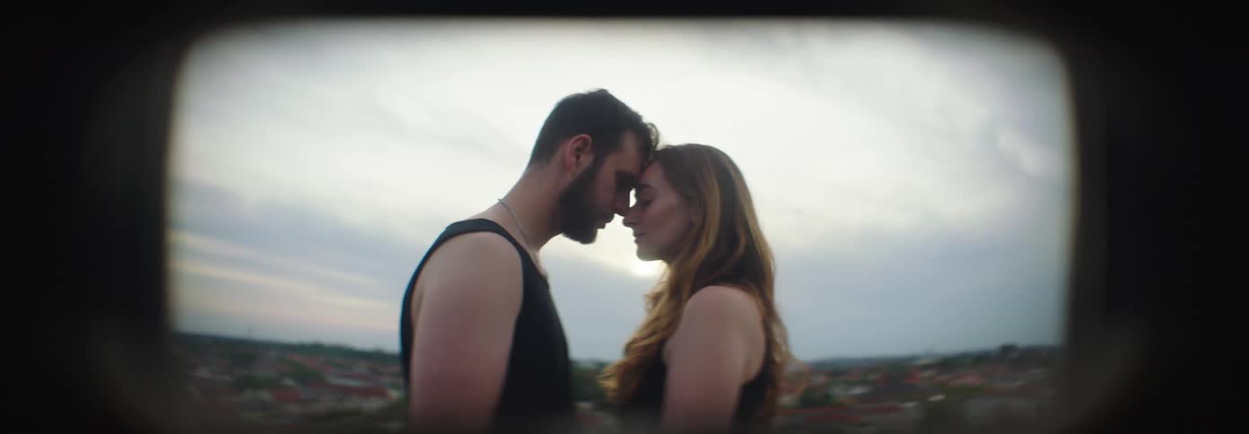Man and woman hold each other with noses touching on a blacony over a red roofed villiage under a dull white clouded sky