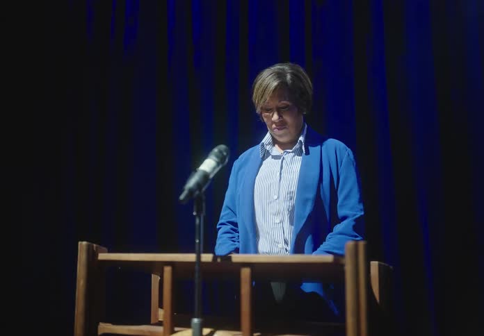 Woman in formal attire speaking at the pulpit with a blue curtain in the background