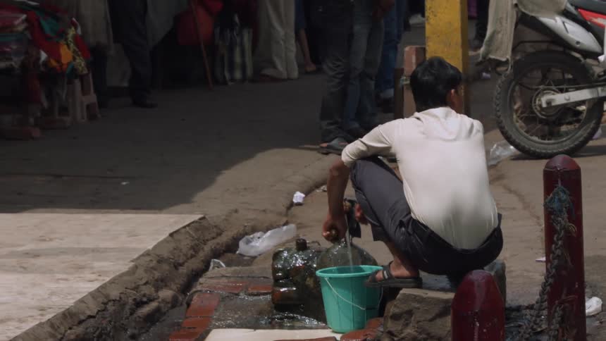 A man is crouching on a street filling a bucket with water