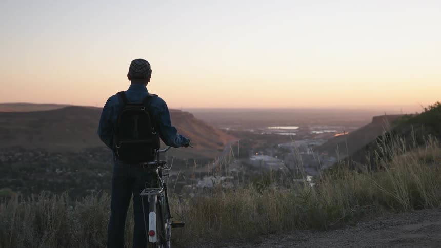 A man stands with his bike on a hill top at dawn