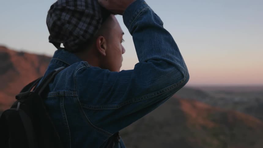 A man adjusts his hat while standing by a mountain