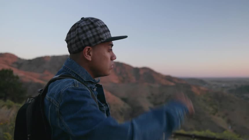 A man adjusts his hat while looking over a mountain range