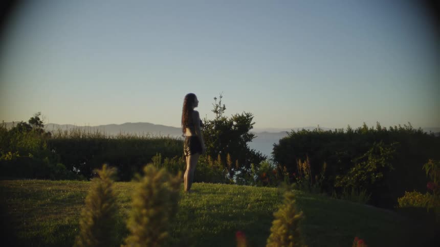 Woman stands on lawn looking over bushes at the water